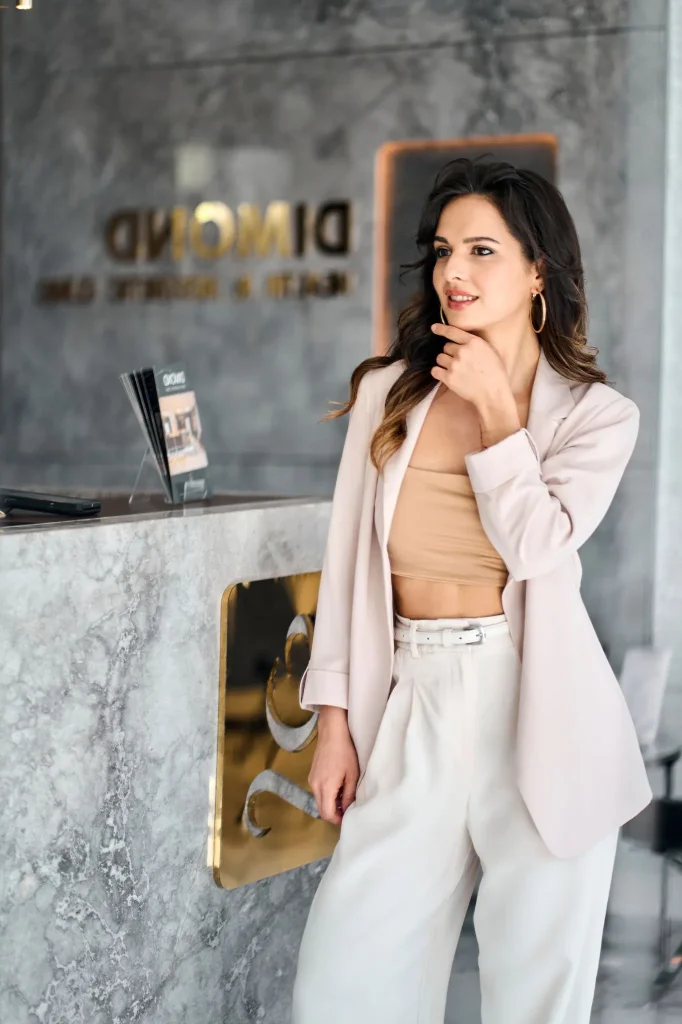 A woman in a stylish beige suit stands thoughtfully in a luxury beauty clinic Dimond Clinic in Sandbanks Road in Poole Dorset, hand on her chick, looking to the mirror. Business Brand Photography Clinic Dorset MS Photography